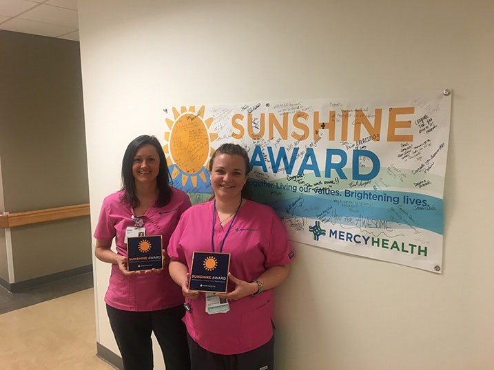 Two women in pink scrubs stand infront of a Sunshine Award sign