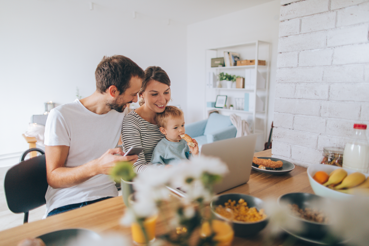 a mother and father with their infant child sit at a kitchen table looking at a laptop
